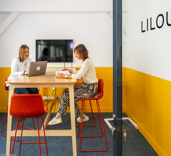 Deux femmes travaillent sur une table haute en bois dans un espace de travail collaboratif aux murs jaunes