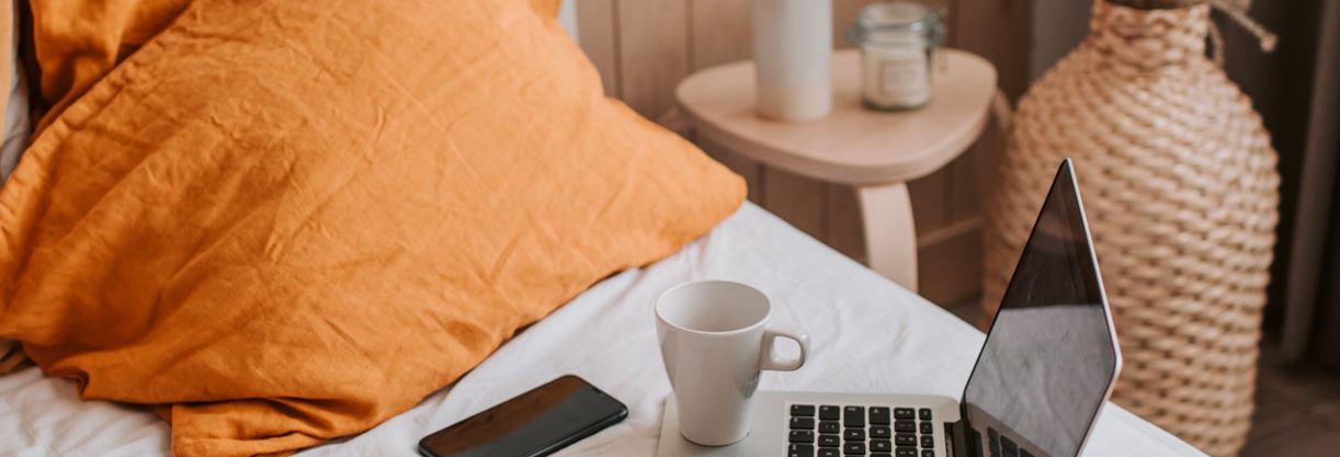 un ordinateur, une tasse, un carnet, un stylo et un téléphone posé sur un lit aux draps blancs et oranges