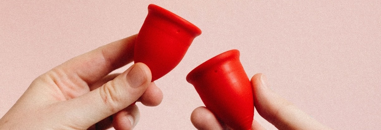 deux mains tiennent des cup menstruelles rouges devant un mur rose