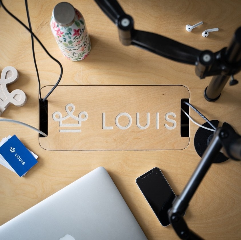 Gravure du logo LOUIS sur un bureau en bois avec un ordinateur, un téléphone, des écouteurs et une gourde à fleurs