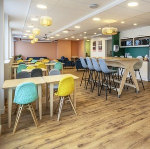 une salle de pause avec cuisine, table haute rectangulaire et plusieurs petites tables avec chaises colorées