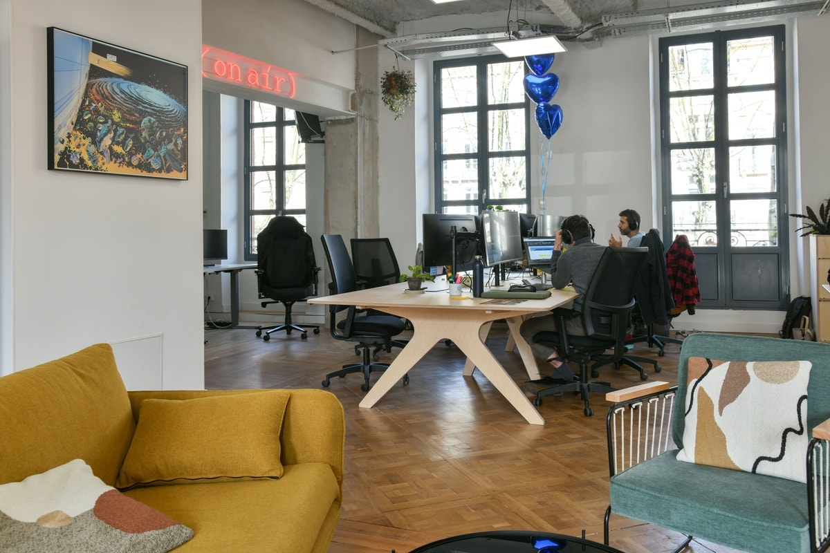 Deux personnes travaillent sur leur ordinateur sur un bureau en bois partagé placé dans un open space avec canapé, fauteuil, néon, table basse.