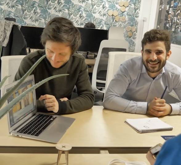 deux personnes souriantes devant une table avec un ordinateur et un carnet posé dessus