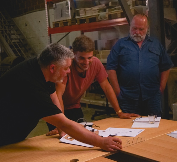 trois personnes regardent les dessins d’une table qui sont posés sur un bureau en bois