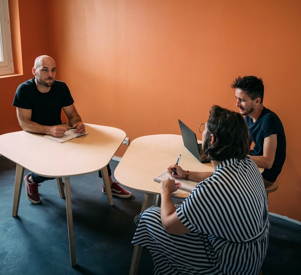 trois salariés discutent assis devant des tables en bois polygonales dans une salle de réunion orange