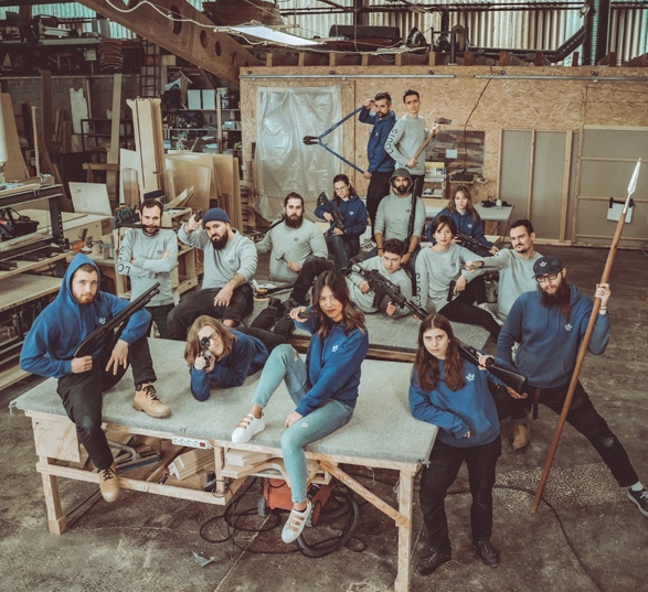 une vingtaine de personnes vêtues de pulls bleus et gris portent des armes en plastique dans un atelier de fabrication de meubles en bois