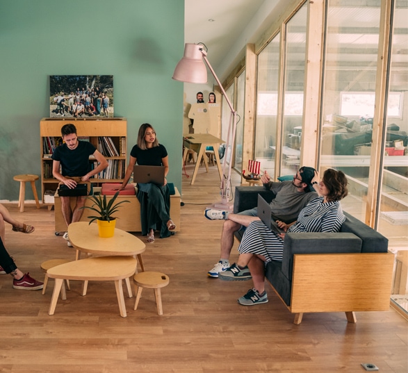 Quatre personnes en réunion dans un espace informel aménagé de deux tables basses, de deux canapés, d’une plante verte, d’une bibliothèque et d’une lampe sur pied.