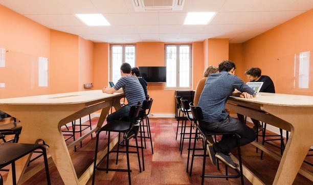 5 personnes travaillent assises sur des chaises hautes devant des tables hautes en bois dans une salle orange