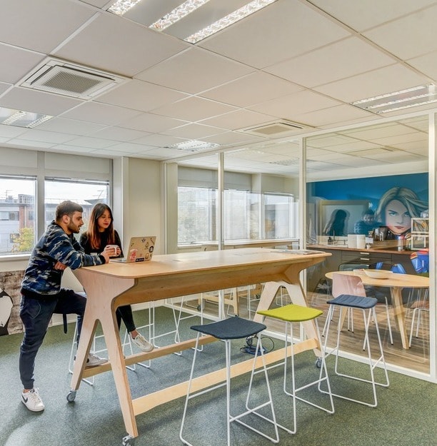 deux personnes travaillent sur une table de réunion rectangulaire haute avec des tabourets hauts colorés dans un espace de travail hybride