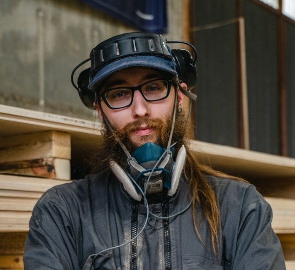 Homme en combinaison de travail grise portant une casquette et un masque de protection dans une usine de production de mobilier en bois