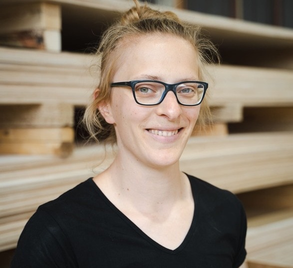 Femme blonde avec des lunettes portant un tee-shirt noir dans une usine de fabrication de meuble en bois