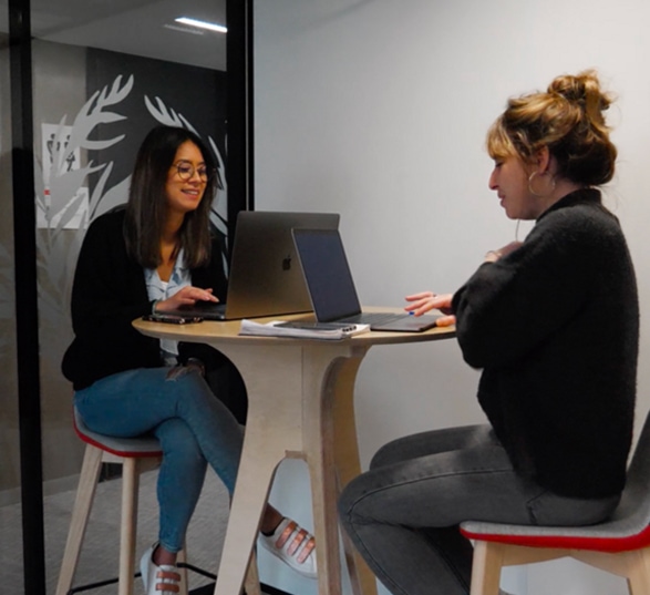 deux femmes font une réunion dans une petite salle et elles sont assise sur des chaises hautes devant une table ronde en bois