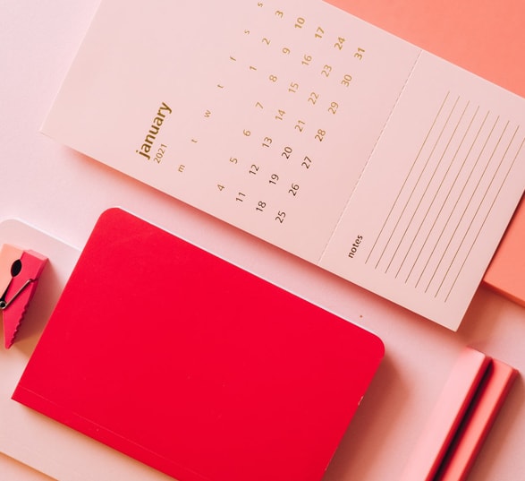 Un calendrier blanc de janvier 2021 posé sur un fond rose à côté d'un carnet rouge et d'une pince à linge rose