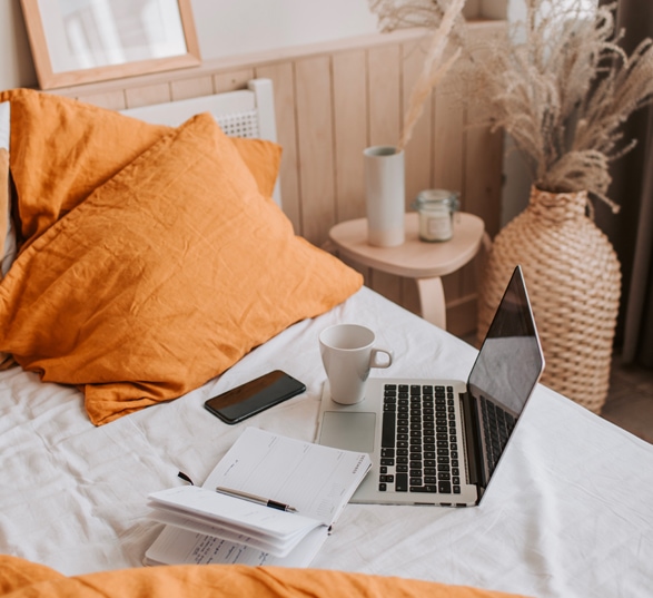 un ordinateur, une tasse et un carnet posé sur un lit blanc avec un oreiller orange