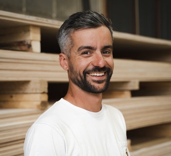 Homme brun avec une barbe portant un tee shirt blanc sur un fond de bois