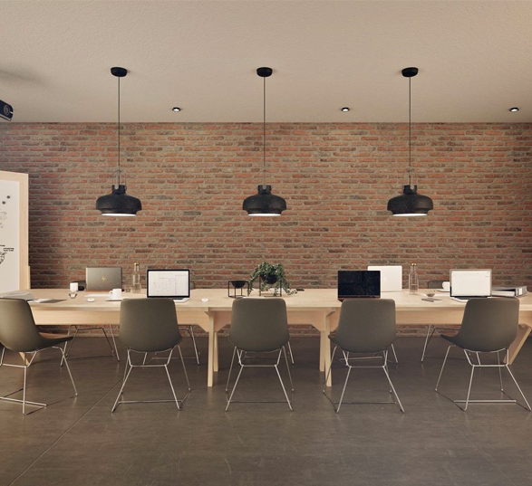 une grande table de réunion en bois clair avec une dizaine de siège de réunion, un mur en brique et des luminaires noirs