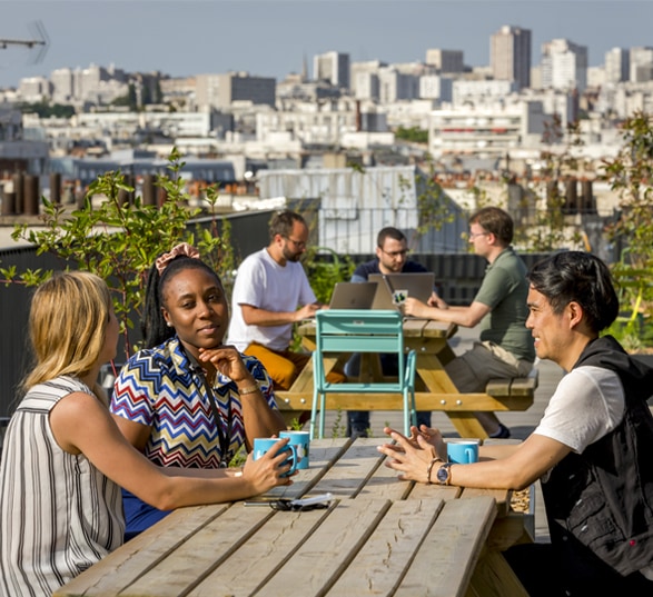 trois personnes discutent sur une table en bois sur un rooftop avec vue sur la ville