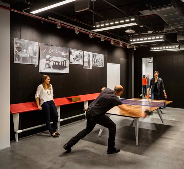 trois personnes sont dans une salle aux murs noirs et deux d'entres-elles font une partie de ping pong