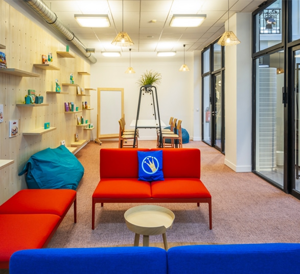 Espace informel en entreprise avec des murs blancs, des canapés rouges, un pouf bleu et du mobilier en bois